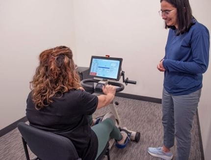 Patient Mandy Ensman on an exercise bike with someone watching her.