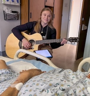 Thumbnail image of a young woman playing guitar for a patient at bedside.