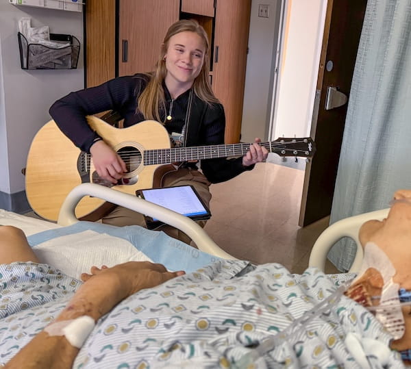 A young, smiling music therapist plays guitar for an elderly patient at the bedside.