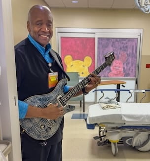 Thumbnail image of a man holding his electric guitar in a patient room.